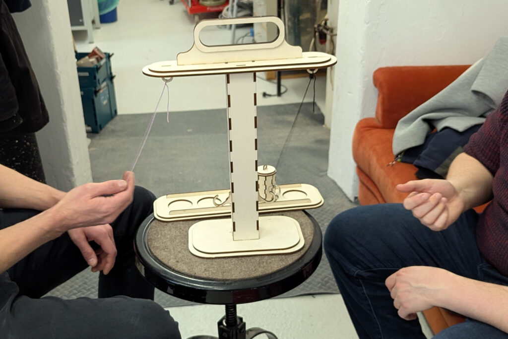 Zwei Personen sitzen sich gegenüber und spielen "Ringing the Bull". Der linke Spieler hält den Ring in der Hand und bereitet seinen nächsten Wurf vor. Der Ring des rechten Spielers prallt gerade vom Haken ab.